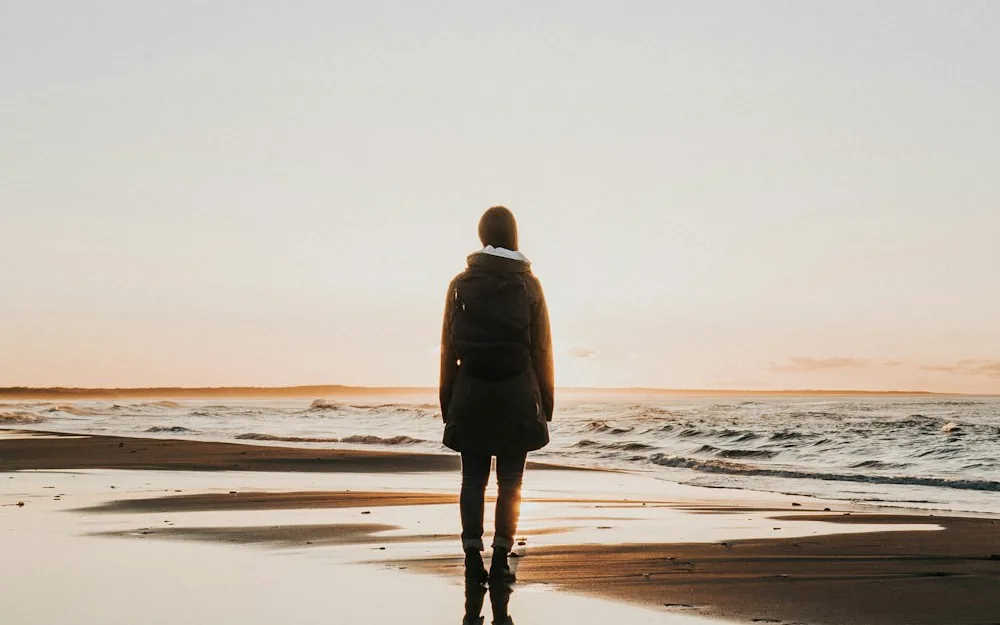 A lone traveler standing at the water's edge, facing the sunset